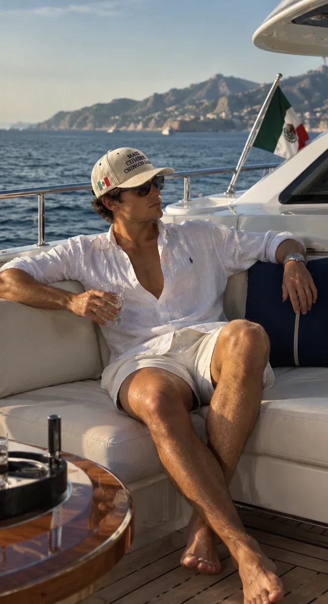 Hombre con la gorra Optimates y camisa de lino blanca, sentado en la cubierta de un yate con la bandera de México al fondo